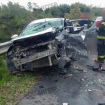 Carro destruído após batida de frente na Rodovia Mogi-Bertioga, com bombeiros no atendimento